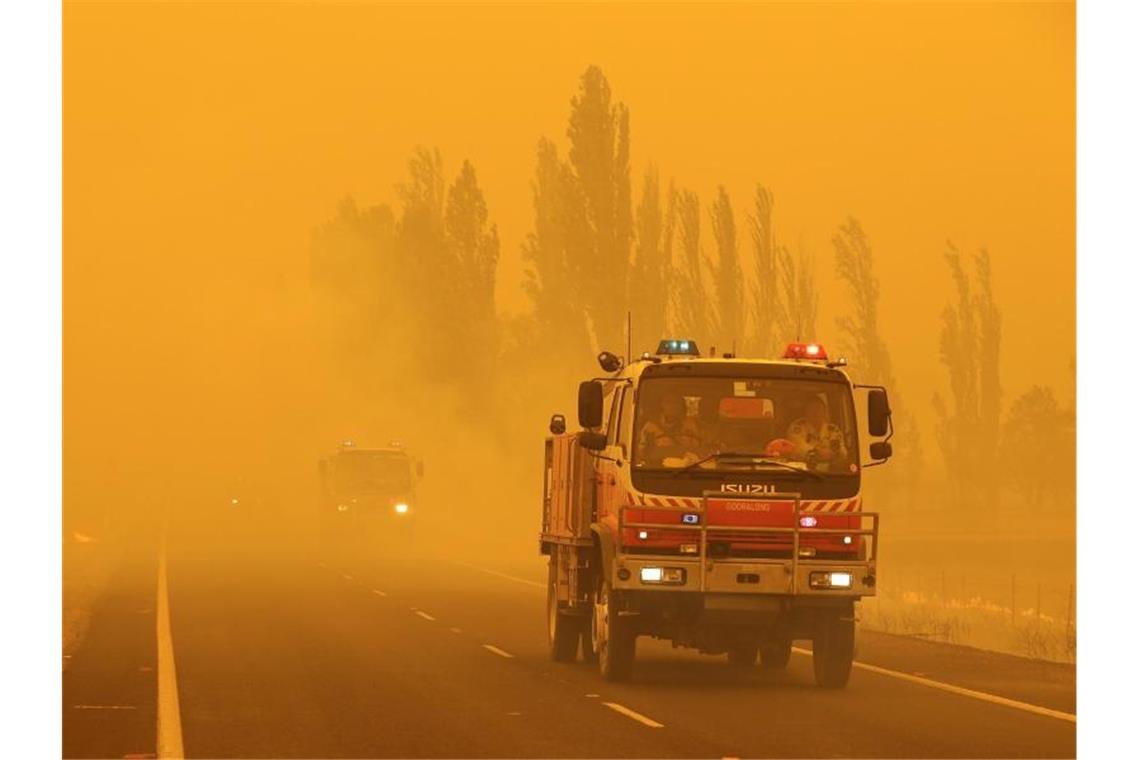 Feuerwehrfahrzeuge fahren in dichtem Rauch in der Region Canberra auf einer Straße, an deren Seiten das Gras brennt. Foto: Rick Rycroft/AP/dpa