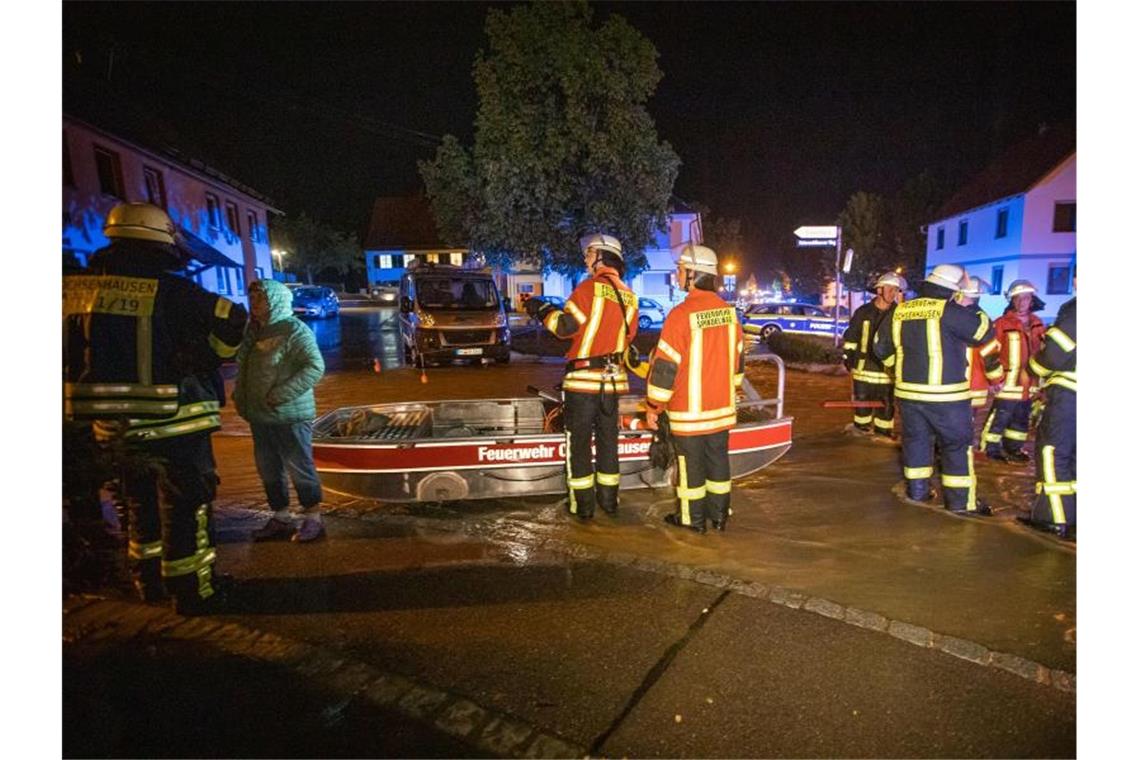 Feuerwehrleute im Kreis Biberach stehen neben einem Feuerwehrboot auf einer Straße in den Fluten eines heftigen Unwetters. Foto: Simon Adomat/VMD Images/dpa