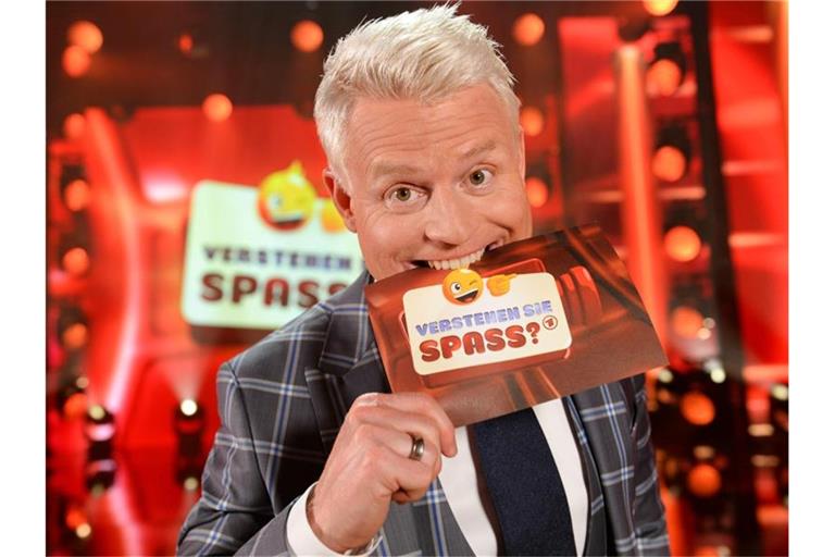 Guido Cantz, Moderator von „Verstehen Sie Spaß?“, steht in der Studiokulisse und beißt auf eine Moderationskarte, auf der das Logo der Sendung abgedruckt ist. Foto: Wolfgang Breiteneicher/SWR/Archiv