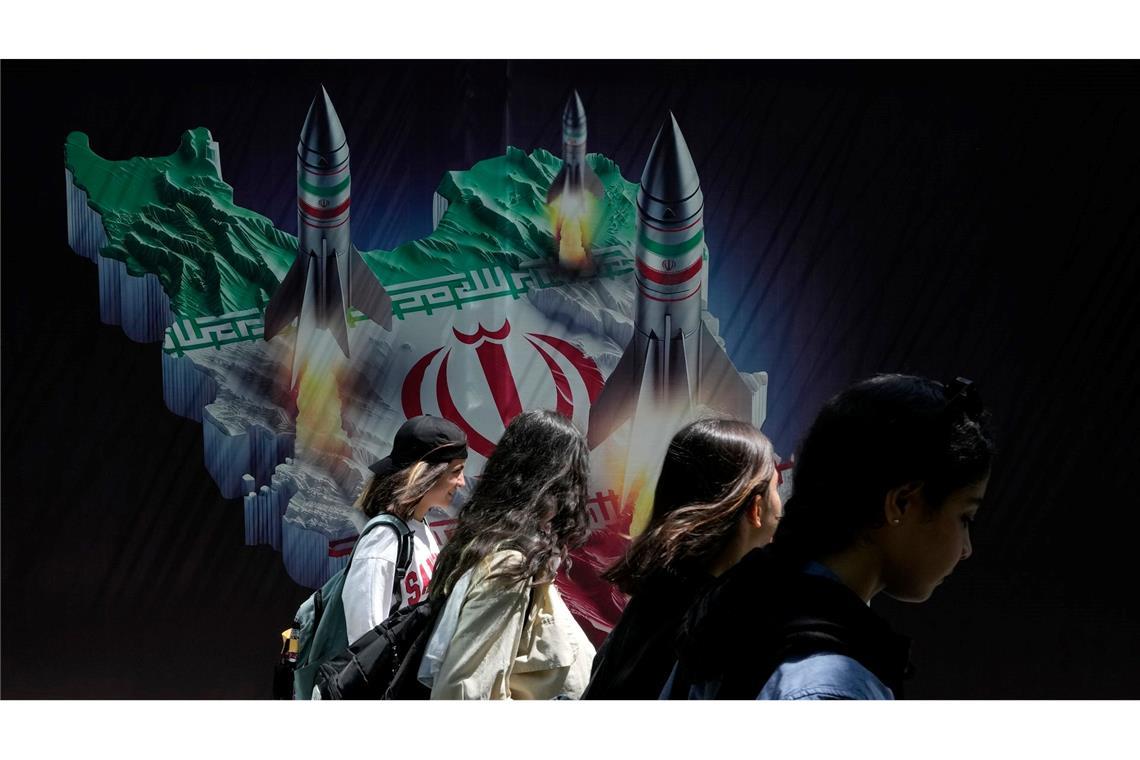 Junge Iranerinnen, die ihr obligatorisches islamisches Kopftuch nicht tragen, gehen in Teheran an einem Banner vorbei, das den Abschuss von Raketen auf einer iranischen Landkarte zeigt.