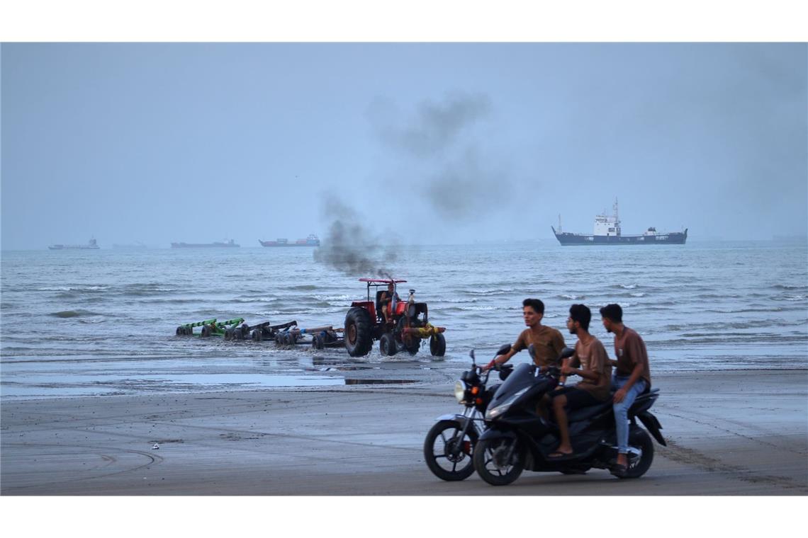 Junge Männer fahren mit Motorrädern am Ufer entlang, während ein Traktor Geräte aus dem Wasser zieht und Handelsschiffe in der Straße von Hormuz vor Anker liegen. (zu dpa: «Rund 400 Schiffe stecken noch im Persischen Golf fest»)