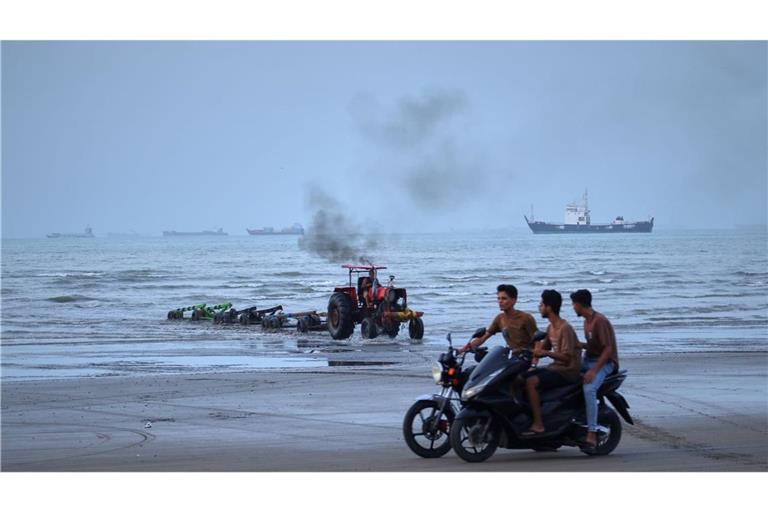 Junge Männer fahren mit Motorrädern am Ufer entlang, während ein Traktor Geräte aus dem Wasser zieht und Handelsschiffe in der Straße von Hormuz vor Anker liegen. (zu dpa: «Rund 400 Schiffe stecken noch im Persischen Golf fest»)