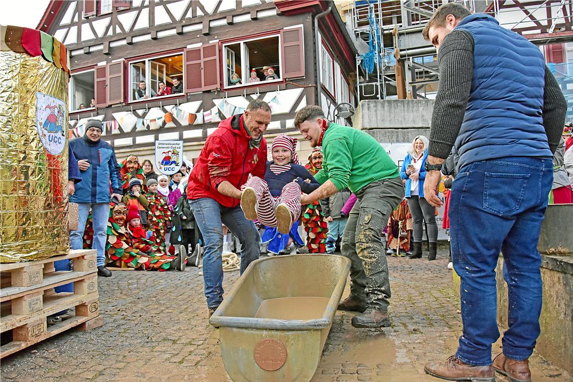 Leonie Heidebrecht lässt sich von so einem närrischen Taufritual kein bisschen die Laune verderben. Bevor sie in die Schlammwanne gesetzt wird, schenkt sie dem Fotografen und der Runde noch ein Lächeln. Auch Bürgermeister Daniel Bogner (rechts) legt mit Hand an und hilft später beim Übergießen mit einer Runde erdigem Wasser-Ton-Gemisch. Fotos: Tobias Sellmaier