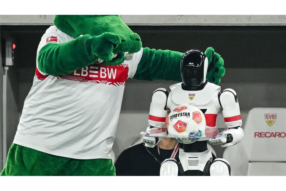 Maskottchen Fritzle neckt einen Roboter mit dem Spielball am Spielfeldrand.