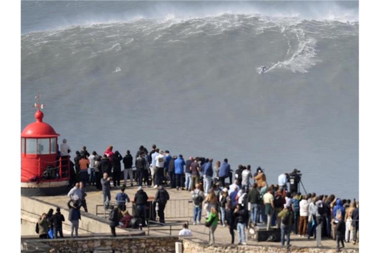 Menschen am Leuchtturm bei Nazaré beobachten einen Surfer am Praia do Norte, der eine Riesenwelle surft. Foto: Armando Franca/AP/dpa