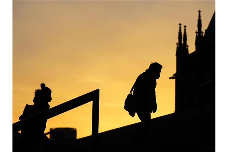 Menschen, die Schutzmasken tragen, gehen im Morgenlicht eine Treppe in Köln hinauf. Die sogenannte Sieben-Tage-Inzidenz in Nordrhein-Westfalen ist aktuell auf 61,7 gestiegen. Foto: Oliver Berg/dpa