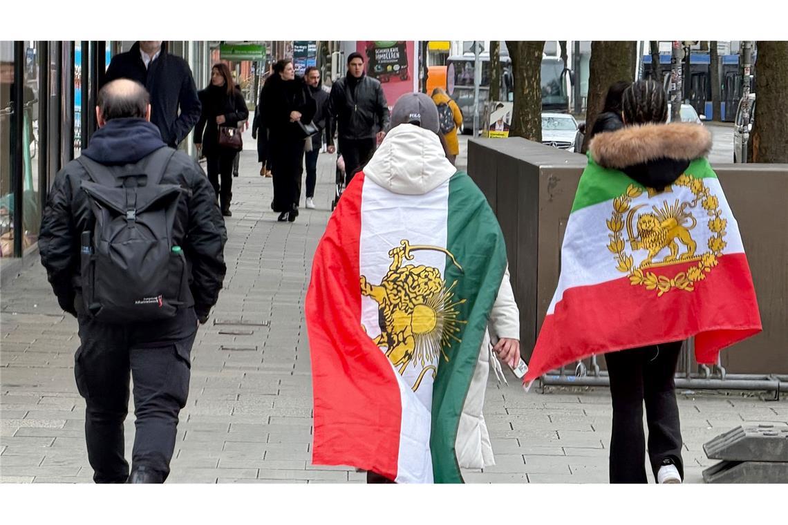 Menschen, die verschiedene ehemalige Flaggen des Iran umhaben, gehen durch die Münchner Innenstadt.