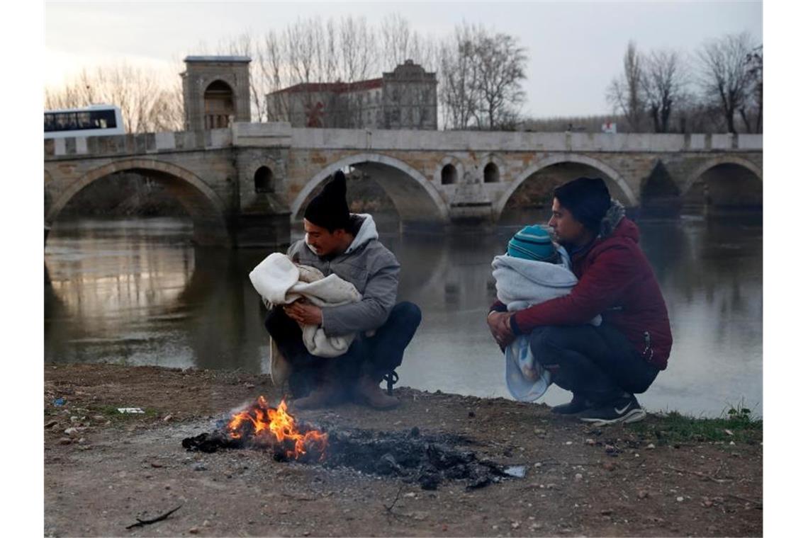 Migranten halten ihre Babys in den Armen neben einem Lagerfeuer am Ufer des Flusses nahe der türkisch-griechischen Grenze. Foto: Darko Bandic/AP/dpa