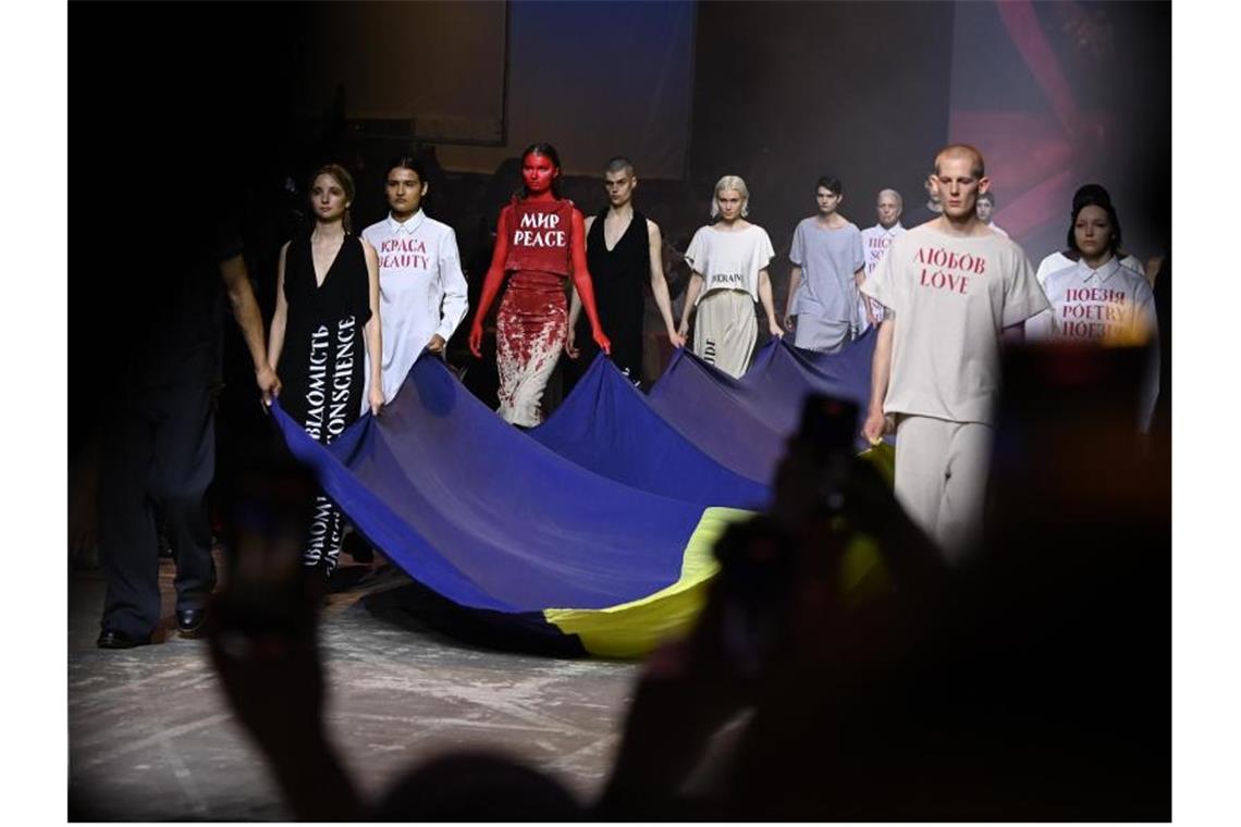 Blutig-rote Models bei Show von ukrainischem Designer Models tragen eine ukrainische Fahne über den Catwalk. „Heute ist nicht die Zeit, über Mode zu sprechen, sondern durch Mode“, so der Designer in einer Videobotschaft. Foto: Jens Kalaene/dpa-Zentralbild/dpa