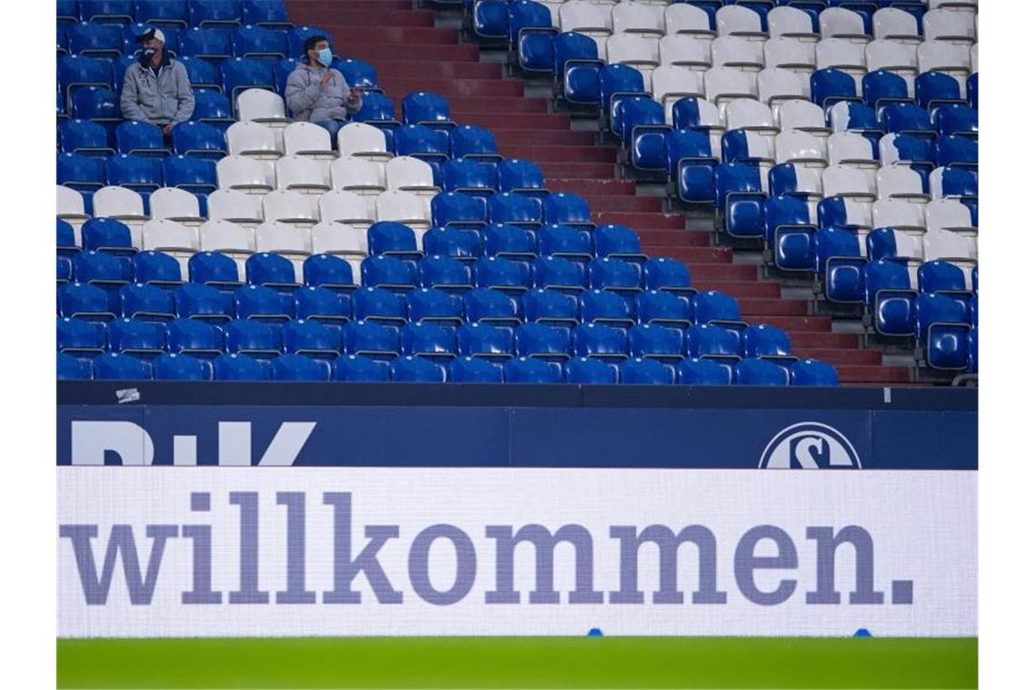 Oberhalb einer Werbebande auf der „Willkommen“ steht, sitzen zwei Schalke Fans. Foto: Guido Kirchner/dpa/Archivbild