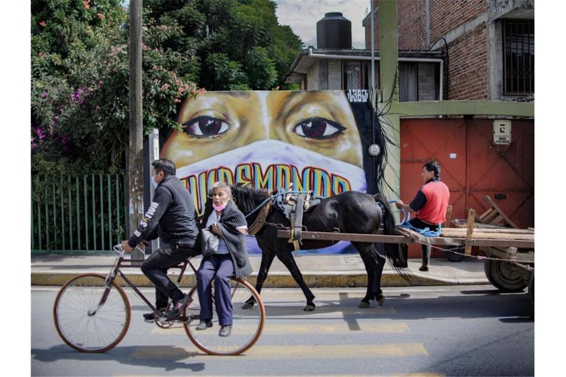 Radfahrer und ein Pferdekarren fahren im mexikanischen Tepetitla an einer Wandmalerei vorbei, die die Bevölkerung zum Maskentragen gegen eine Corona-Infektion auffordert. Foto: Jesus Alvarado/dpa