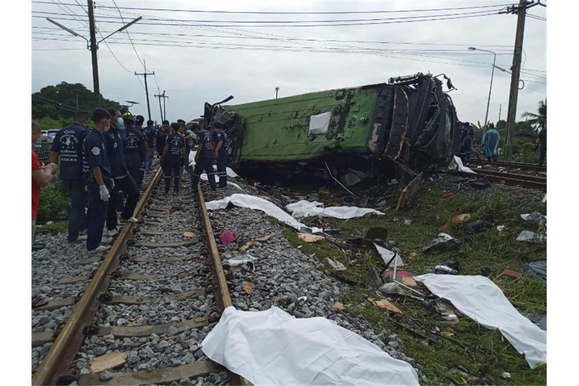 Zug stößt in Thailand mit Bus zusammen - mindestens 17 Tote Rettungskräfte stehen an einem beschädigten Zug und neben mit weißen Tüchern bedeckten Leichen. Mindestens 17 Menschen sind am Sonntag in Thailand beim Zusammenstoß eines Busses mit einem Zug ums Leben gekommen. Foto: Uncredited/Daily News/AP/dpa