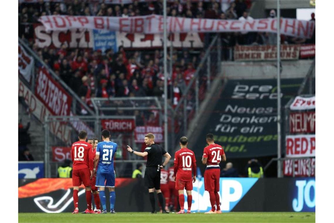 Schiedsrichter Christian Dingert (M) spricht mit Spielern, während Fans auf der Tribüne ein beleidigendes Banner zeigen. Foto: Michael Probst/AP/dpa