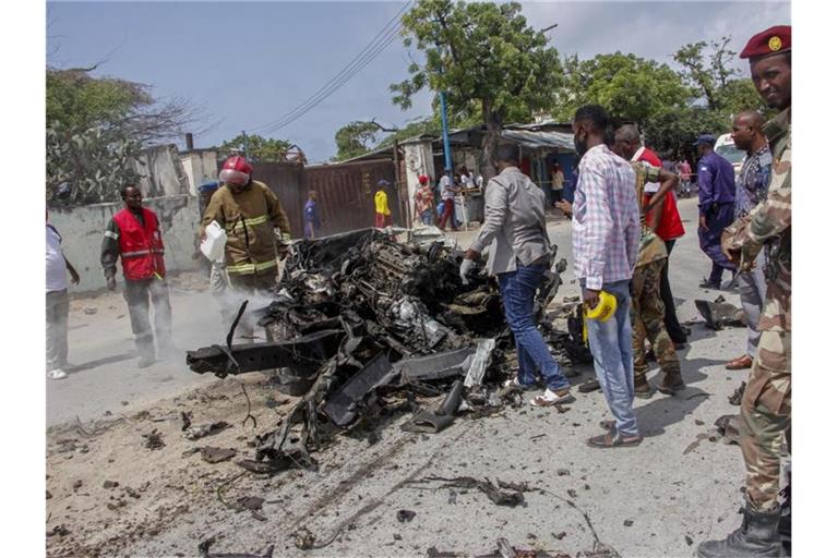 Sicherheitskräfte und Zivilisten stehen in der Nähe eines Autowracks nach dem ersten Anschlag im somalischen Mogadischu. Die islamistische Terrormiliz Al-Shabaab hat nun sechs weitere Mörsergeschosse abgefeuert. Foto: Farah Abdi Warsameh/AP/dpa