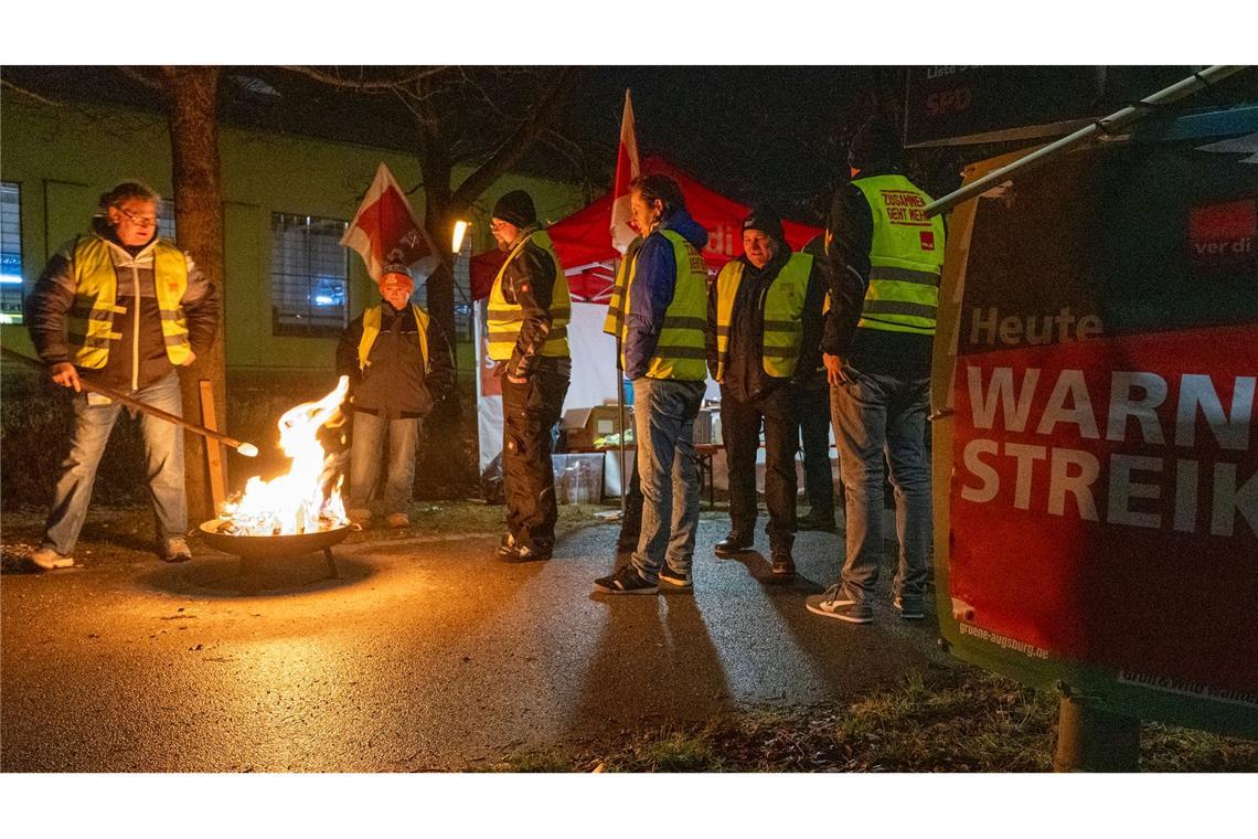 Streikende in Warnwesten wärmen sich in Augsburg an einem Feuer während eines Warnstreiks im ÖPNV.