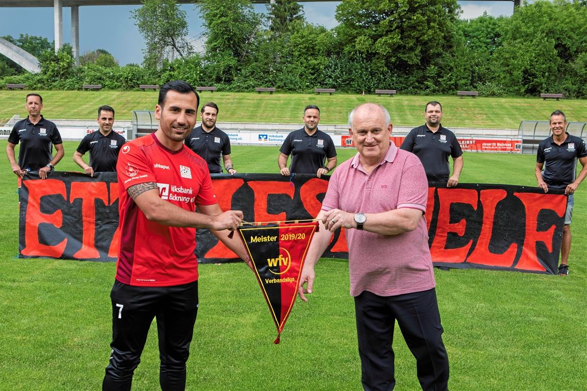 TSG-Kapitän Oguzhan Biyik nimmt den Meisterwimpel vom WFV-Verbandsspielausschuss-Vorsitzenden Harald Müller entgegen. Im Hintergrund freuen sich der sportliche Leiter Marco Erdmann, Teammanager Isaak Avramidis, Co-Trainer Salvatore Rivarolo, Trainer Evangelos Sbonias, Co-Trainer Darko Milosevic und Vorstand Sport Rüdiger Lüftner (hinten von links). Foto: A. Becher