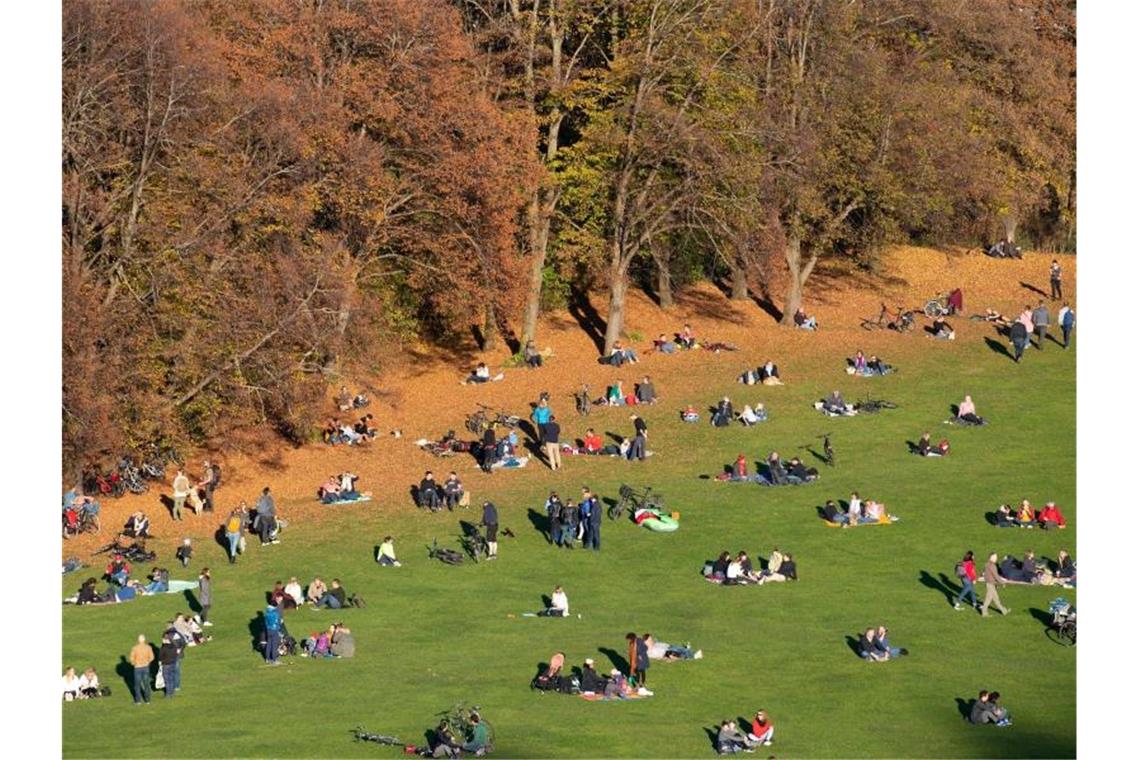 Zahlreiche Menschen sitzen im Münchner Olympiapark vor herbstlich verfärbten Bäumen auf einer Wiese. Foto: Sven Hoppe/dpa