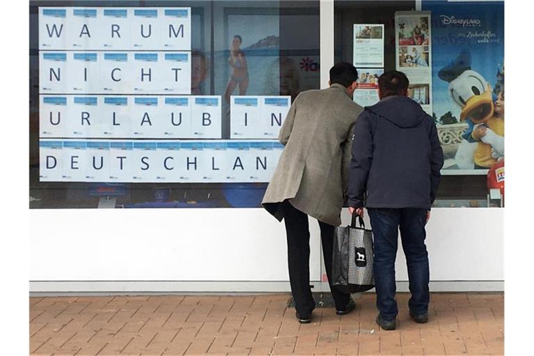 Zwei Männer schauen in das Schaufenster eines Reisebüros, an dem mit großen Buchstaben „Warum nicht Urlaub in Deutschland“ zu lesen ist. Foto: Bernd Thissen/dpa
