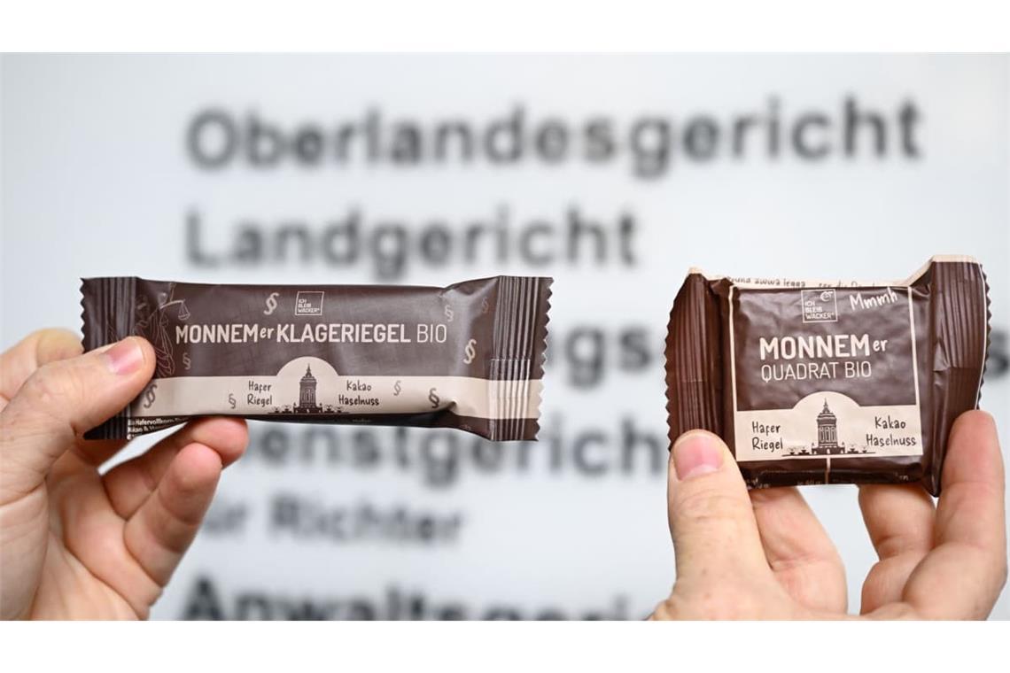 Zwei Riegel der Firma Wacker werden vor dem Eingangsschild des Oberlandesgerichts in die Höhe gehalten. Ritter Sport sieht durch einen quadratischen Riegel der Mannheimer Firma Wacker seine Marke verletzt und klagt.