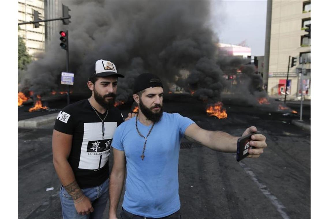 Zwei Teilnehmer einer Demonstration fotografieren sich vor einer brennenden Straßenblockade mit einem Smartphone. Foto: Hassan Ammar/AP/dpa