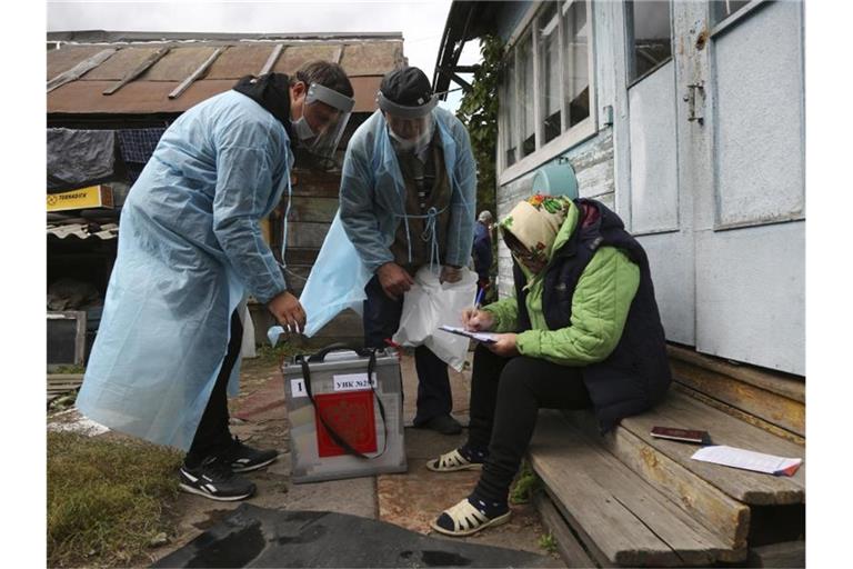 Zwei Wahlhelfer stehen neben einer Frau, die auf den Stufen vor einem Haus sitzt und Dokumente ausfüllt. Foto: Evgeniy Sofiychuk/AP/dpa